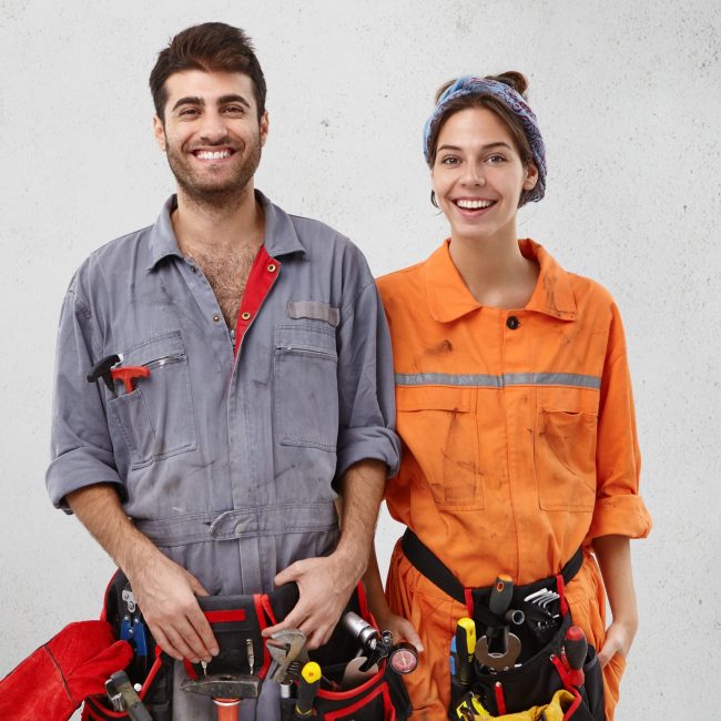 Heureux bricoleur barbu et sa compagne se tiennent l'un à côté de l'autre, se sentent soutenus et aidés, travaillent en équipe amicale, remodèlent des objets, sourient joyeusement, isolés sur un mur de studio en béton blanc